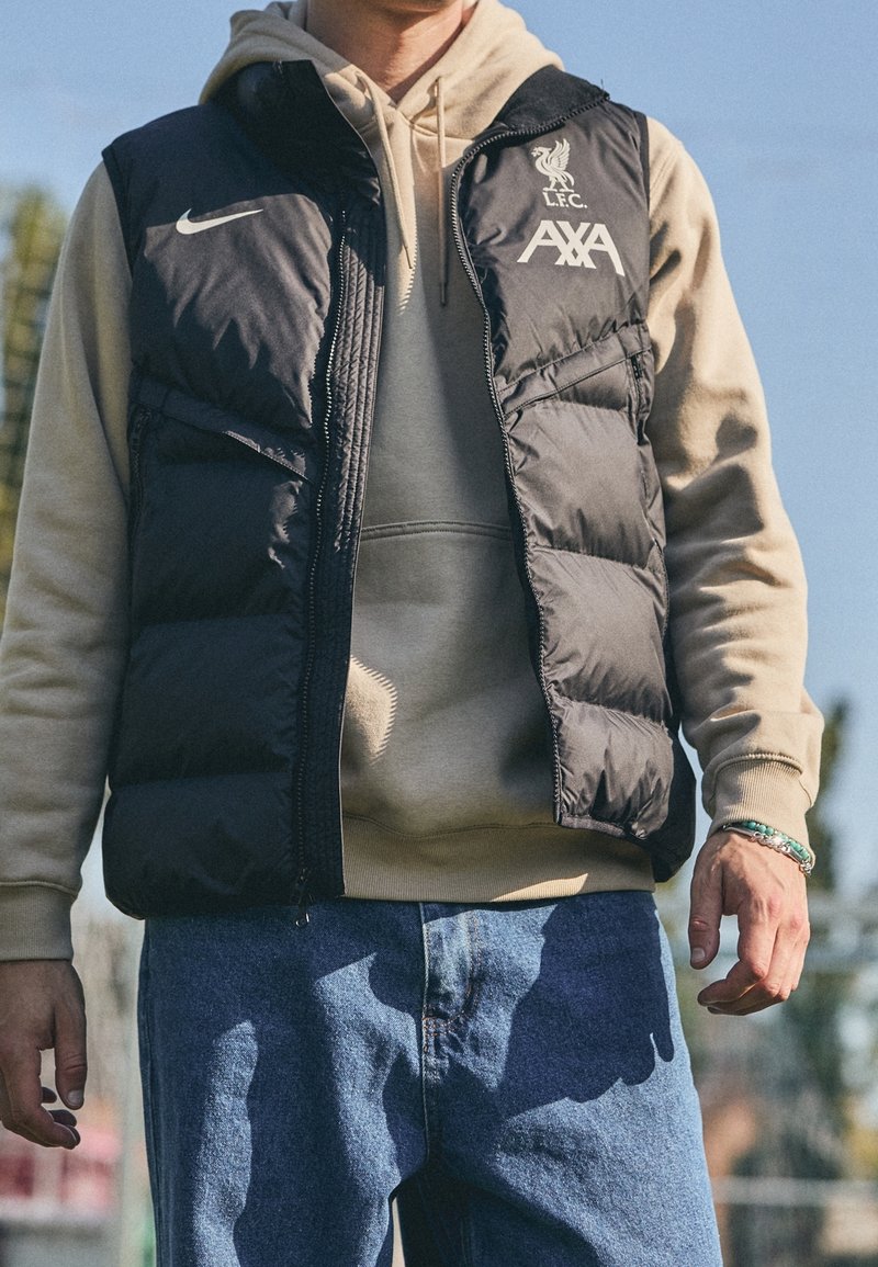 Homme portant un sweat à capuche beige sous une doudoune noire Nike avec le logo LFC AXA et un jean bleu, debout à l'extérieur sous un ciel clair.