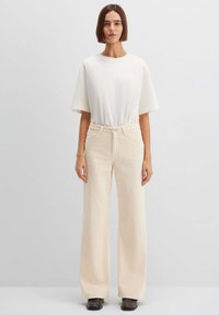 Beige Cord-Hosen mit weitem Bein, vorne Taschen, Knopfverschluss und einer weichen, strukturierten Oberfläche, kombiniert mit einem schlichten weißen T-Shirt.