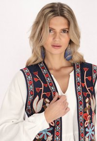 Jūras spēku izšūta vests ar ziedu rakstiem sarkanā un zilā krāsā. Zem tā valkāta balta blūze. Nozīmīgi ģeometriski akcenti gar malām.