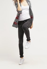 Grå mönstrad kofta med röda detaljer över en vit linne, kombinerat med svarta jeans och vita sneakers, vilket visar en avslappnad stil.