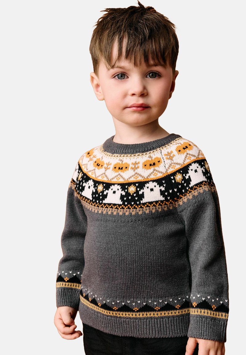 Ung dreng med kort brunt hår iført en grå sweater med orange og hvide Halloween-tema mønstre inklusive spøgelser og græskar.