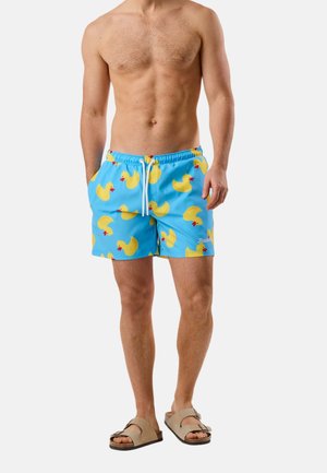 Oberkörperfreier Mann trägt blaue Badeshorts mit gelbem Gummientenmuster und beige Sandalen, steht vor einem schlichten Hintergrund.