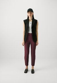 Crni bezrukavni sako preko bijele uske majice, uparen s burgundskim tapered hlačama i crnim mokasinkama. Model nosi crnu kapu.