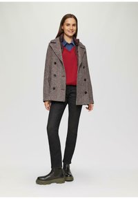 Grijs dubbele jas met visgraatpatroon en donkere knopen. Draagt over een rode trui en een blauwe blouse, gecombineerd met zwarte jeans en donkere laarzen.