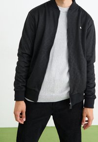 Black bomber jacket with a subtle embossed pattern, front zipper, and ribbed cuffs, worn over a light gray ribbed sweater, paired with black pants.