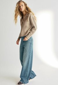 Cardigan tricotat bej cu fermoar, având o buzunare și o literă brodată. Asortat cu blugi cu picioare largi de culoare albastru deschis și pantofi închiși la culoare.