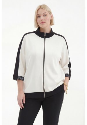 Jeune femme aux cheveux blonds courts portant une veste blanche zippée avec des manches noires et un pantalon noir, debout devant un fond blanc.