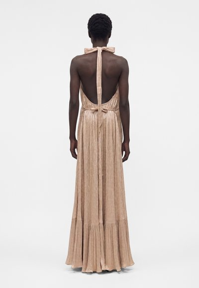 Άτομο που φοράει αμάνικο, λαμπερό μπεζ φόρεμα με halter λαιμόκοψη, ανοιχτή πλάτη και ζώνη στη μέση, στέκεται μπροστά σε απλό λευκό φόντο.