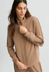 Bruine sportieve zip-up jas met hoge kraag, zijzakken en gaasaccenten op de mouwen, gemaakt van een zachte, gestructureerde stof.