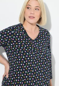 Frau mit kurzem blonden Haar, die ein dunkelblaues Oberteil mit kleinen Teddybären, rosa Tassen und weißen kreisförmigen Mustern trägt und zur Seite schaut.