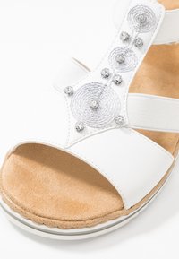 Sandale blanche avec des accents spirales en argent et des strass, semelle intérieure beige douce et sangles élastiques pour un maintien sécurisé. Semelle extérieure texturée.