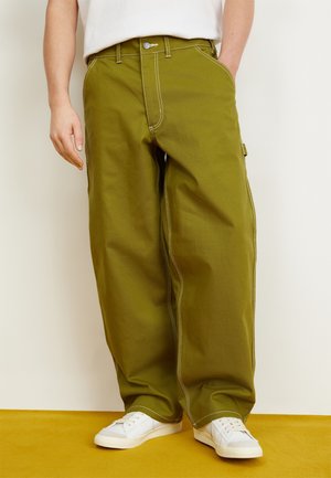 Personne portant un pantalon ample vert olive avec des coutures blanches, des baskets blanches, se tenant sur un sol jaune contre un mur blanc.