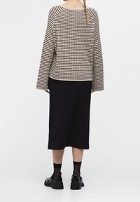 Vrouw met een lange vlecht, gekleed in een gestreepte beige en zwarte trui met een textuur, zwarte midi-rok, doorschijnende sokken en zwarte chunky schoenen, staande met de rug naar ons toe.