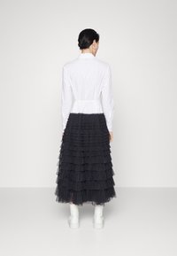 Camicia bianca a maniche lunghe con vita arricciata, abbinata a una gonna in tulle nero a strati. La modella indossa stivaletti bianchi chunky.