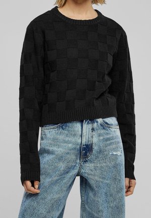 Henkilö, joka käyttää teksturoitua mustaa neulekutoa, jossa on ruutukuvio, ja vaaleansinisiä, korkeavyötäröisiä kulutettuja farkkuja.