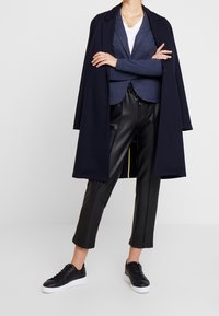 Abrigo largo azul marino sobre un cárdigan azul, camiseta blanca, pantalones de cuero negro y zapatillas negras con suela blanca. Conjunto casual en capas.