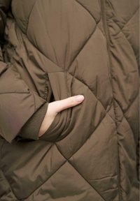 Veste matelassée vert olive avec une poche latérale, dotée de poignets élastiques et d'une texture lisse. Gros plan sur une main atteignant la poche.