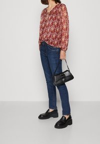 Donna che indossa una blusa a maniche lunghe con motivo, jeans blu, mocassini neri con plateau e tiene una borsa nera con manico a catena.