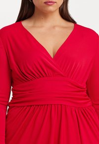 Červené šaty s hlubokým výstřihem do V a dlouhými rukávy. Mají nabíranou látku v pase a splývavou sukni, vyrobené z hladkého, elastického materiálu.