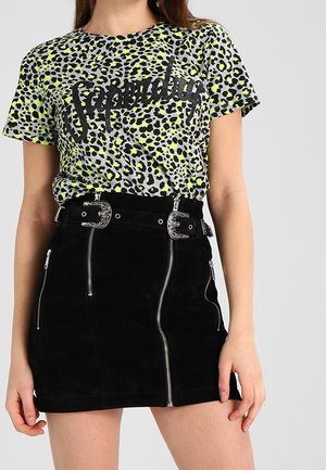 Kobieta w neonowo zielonej i czarnej koszulce w panterkę z napisem "superdry" oraz czarnej mini spódniczce z srebrnymi zamkami błyskawicznymi i paskiem z podwójną klamrą.