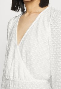 Vestido branco em tecido texturizado, com decote em v profundo e mangas bufantes, apresentando uma cintura franzida e uma silhueta fluida.