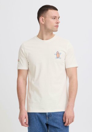 Nuori mies, joka pukeutuu kermanväriseen T-paitaan, jossa on pieni värikäs sarjakuvahahmopainatus vasemmassa rinnassa, yhdistettynä sinisiin farkkuihin, katsoo oikealle.