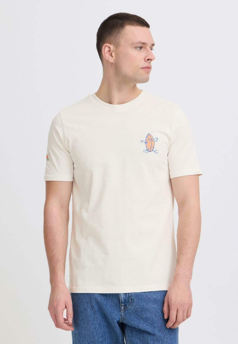 Νεαρός άνδρας φοράει κρεμ μπλουζάκι με μικρό πολύχρωμο σχέδιο κινουμένων σχεδίων στο αριστερό μέρος του στήθους, συνδυασμένο με μπλε τζιν, κοιτάζοντας προς τα δεξιά.