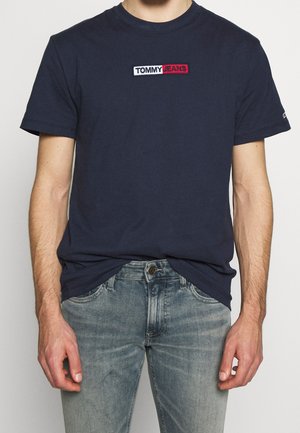Man in een marineblauwe T-shirt met korte mouwen met het "Tommy Jeans" logo en vervaagde blauwe jeans, staande tegen een neutrale achtergrond.