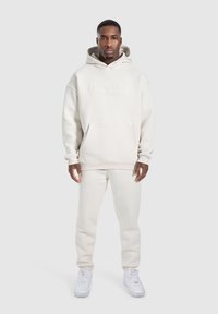 Sudadera con capucha color crema, con un bolsillo delantero y un logo bordado, combinada con pantalones de chándal a juego y zapatillas blancas.