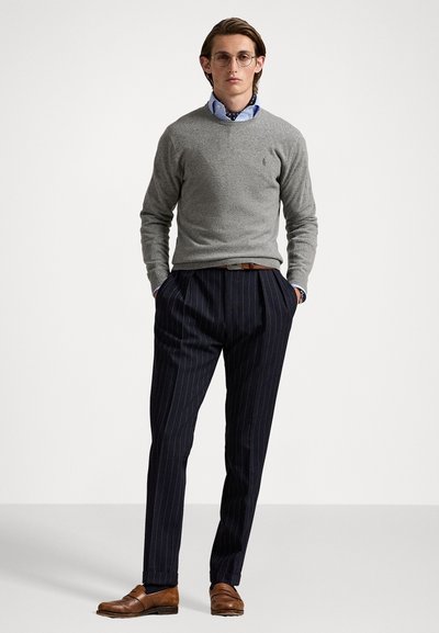 Pull en tricot gris, chemise à col bleu, pantalon rayé bleu marine, mocassins marron. Le mannequin se tient avec les mains dans les poches sur un fond blanc.