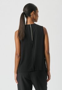 Črn brezrokavnik z visokim ovratnikom in zadrgo na sredini hrbta. Gladek material s sproščenim krojem, v kombinaciji s črnimi hlačami.