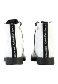 Botas de tobillo de charol blanco con una franja vertical negra que presenta el texto "Calvin Klein Jeans". Suela de goma de contraste; cierre de cremallera en el costado.