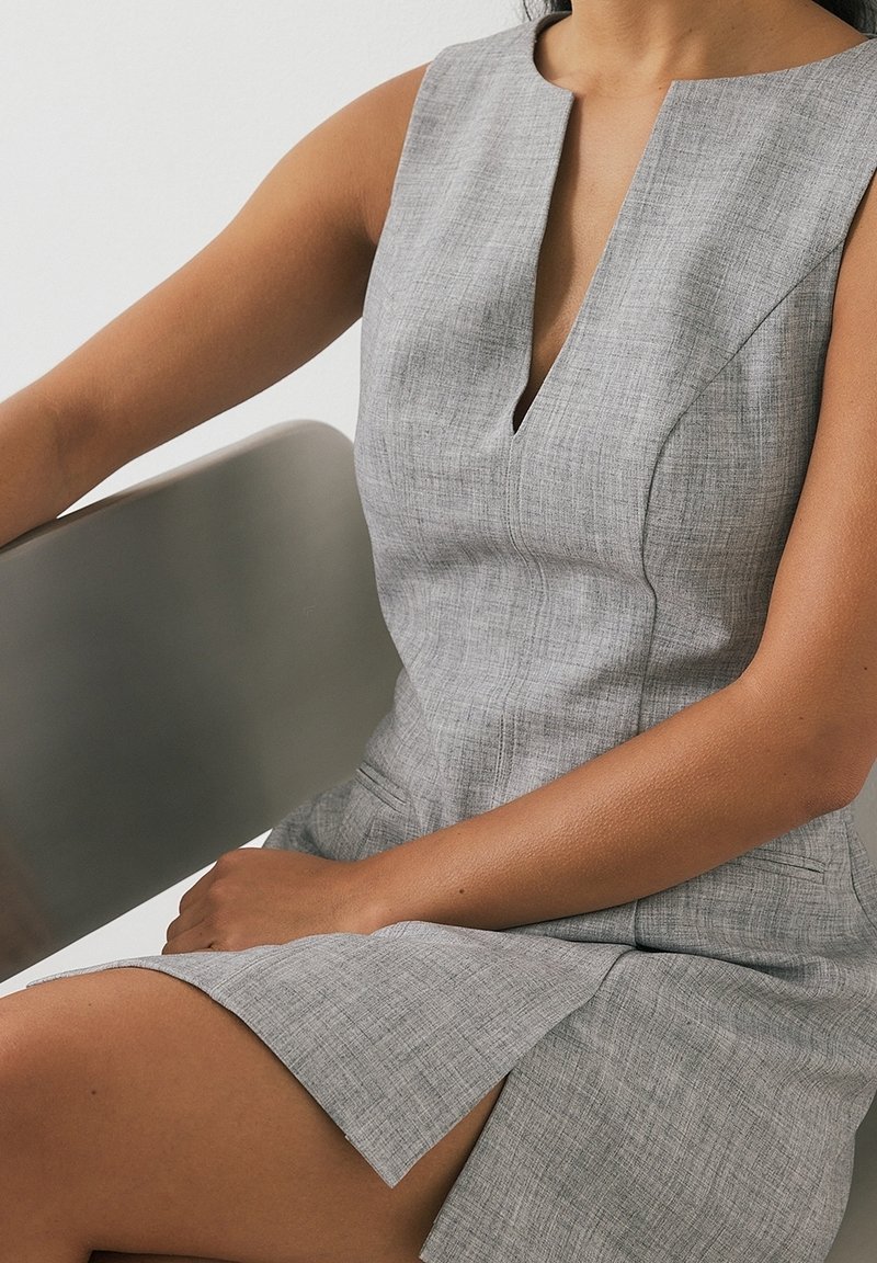 Femme portant une robe grise sans manches avec une fente devant, assise sur une chaise gris clair sur un fond uni.