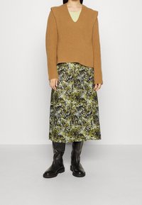 Pull en mailles côtelées marron avec un col en V, superposé sur un haut vert et une jupe midi à motif floral dans des tons de vert et de noir, accompagnée de bottes noires.