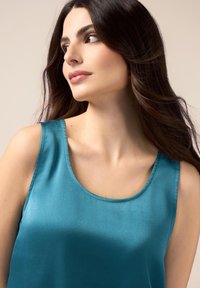 Türkises Satin-Trägertop mit einem runden Ausschnitt und breiten Trägern. Glatte Textur mit sanftem Glanz, die die Schultern und das Schlüsselbein betont.