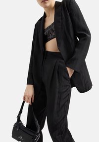 Blazer nero su misura con una texture liscia, abbinato a pantaloni a vita alta. Top corto nero con paillettes visibile sotto. Piccola borsa nera.