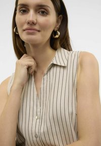Blusa sin mangas en color beige con rayas verticales en azul marino, con cuello y escote en V, que presenta detalles de botones en el frente y una textura suave.