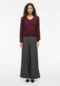 Femme debout, portant un pull en V bordeaux avec des manches texturées, un pantalon gris à jambes larges et des chaussures noires à bout pointu.