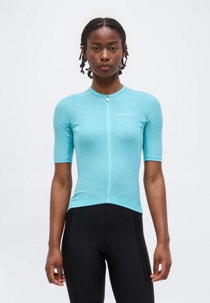 Női modell világoskék, rövid ujjú, testhezálló kerékpáros felsőt visel elöl cipzárral és fekete kerékpáros rövidnadrágot.