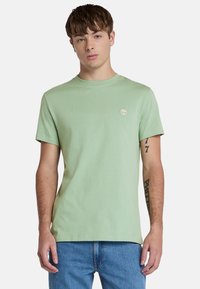 Camiseta de algodón verde claro con cuello redondo, mangas cortas y un pequeño logo en el pecho. Combinada con unos jeans de mezclilla azules.