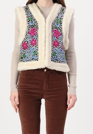 Kobieta w kremowej kamizelce z polaru z kolorową kwiatową haftowaną dekoracją, na beżowym prążkowanym topie z długim rękawem i brązowych spodniach sztruksowych.