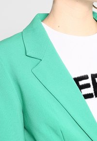 Veste turquoise avec un col cranté, un tissu texturé et une silhouette structurée, associée à un t-shirt blanc avec des inscriptions noires.