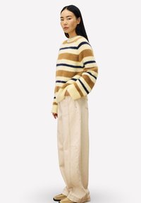 Vrouw die zijwaarts staat, gekleed in een fluffy gestreepte trui in beige, bruin en marineblauw, met brede beige broek en tan schoenen.