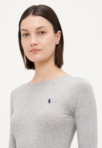 Pilka ilgomis rankovėmis marškinėliai pagaminti iš minkštos medžiagos, ant krūtinės yra mažas mėlynas logotipas. Apykaklė yra apvali ir turi lygią tekstūrą.