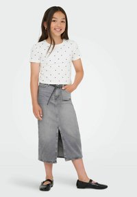 Šypsena mergina stovi vilkėdama baltą trumpą rankovę marškinėlį su širdelėmis, pilką midi džinsinę sijoną su priekiu išpjova ir juodas Mary Jane batelius.