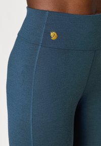 Wysokie, ciemnoniebieskie legginsy z elastycznego materiału, z przeszyciami i małym, żółtym haftowanym logo na pasku.