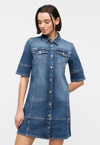 Γυναίκα που φοράει φόρεμα από τζιν με κουμπιά, κοντά μανίκια και τσέπες στο στήθος, κοιτάζοντας προς τα κάτω μπροστά σε ένα απλό φωτεινό φόντο.