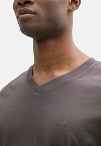 Tricou gri cu decolteu în V, cu mâneci scurte, realizat dintr-un material moale din bumbac. Se remarcă printr-un logo subtil pe piept și o textură netedă.