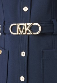 Abito blu navy in tessuto testurizzato, con fibbia in oro con logo MK su una cintura abbinata e bottoni dorati accentati sulla parte anteriore.