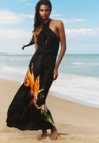 Black halter maxi dress featuring a keyhole cutout and vibrant tropical bird and flower print at the hem; lightweight fabric flows around legs.